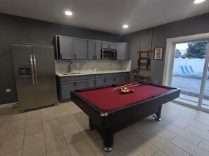 A modern kitchen with grey cabinets, a stainless steel refrigerator, and a pool table with red felt.