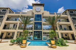 A multi-story hotel with a central atrium, a pool, and palm trees. - New Ground Floor 1BR Condo ~ Close to Eagle Beach (Noord)