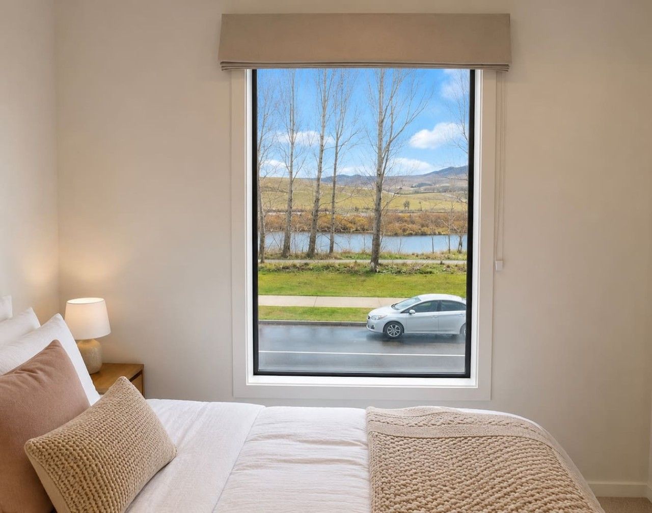 A bedroom with a large window, a bed with white bedding, and a lamp on a bedside table.