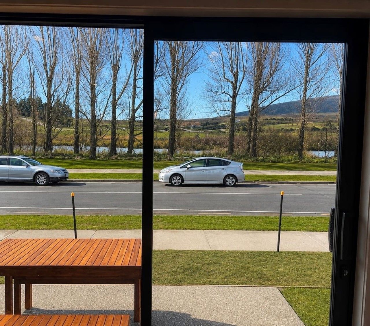 Vista desde el interior de un edificio que muestra una calle con dos autos, un área con césped y árboles sin hojas.
