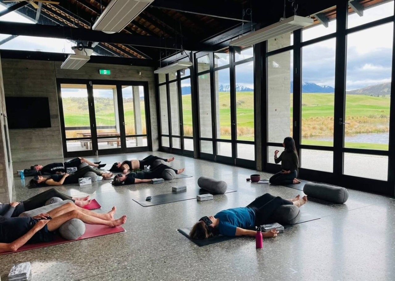 A group of people lying on mats in a spacious room with large windows and a view of a green landscape.