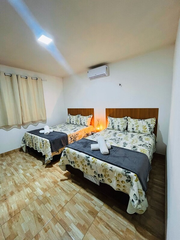 A hotel room with two beds, a window with curtains, an air conditioning unit, and a tiled floor.