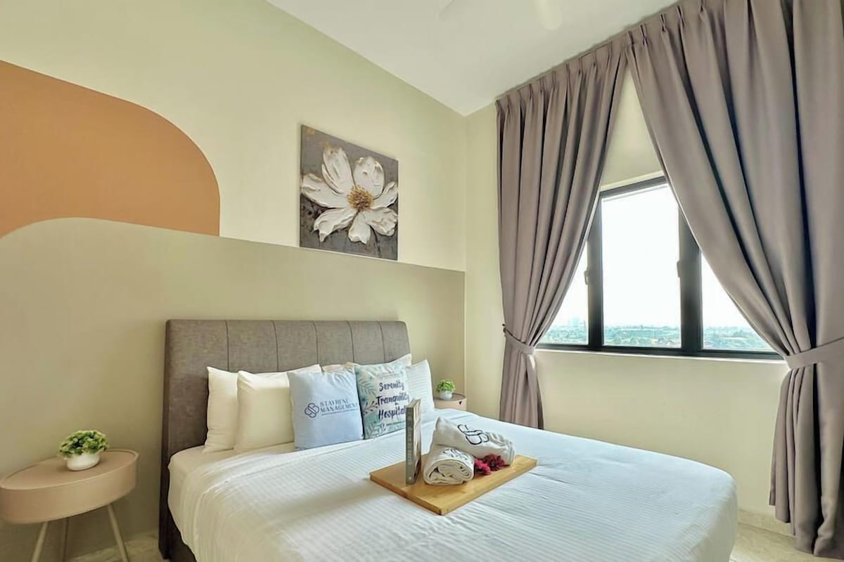A hotel room with a large bed, a window with curtains, a wall-mounted flower painting, and a bedside table with a plant.