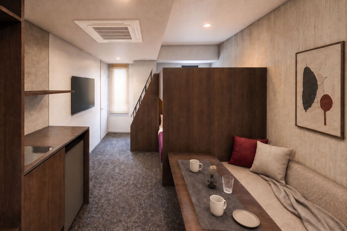 A compact hotel room with a kitchenette, a sofa, a dining table, and a TV mounted on the wall.