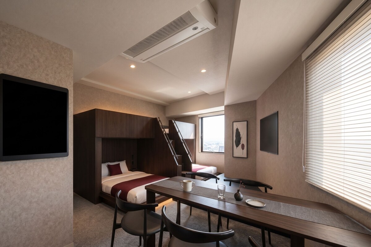 A hotel room with a bed, a desk, chairs, a window with blinds, and a wall-mounted air conditioning unit.