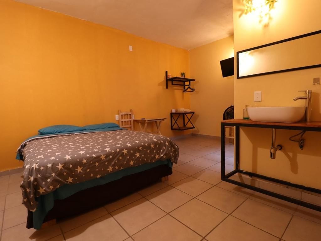 Una habitación con una cama, una mesita, una silla, una estantería y un baño con lavabo y espejo.