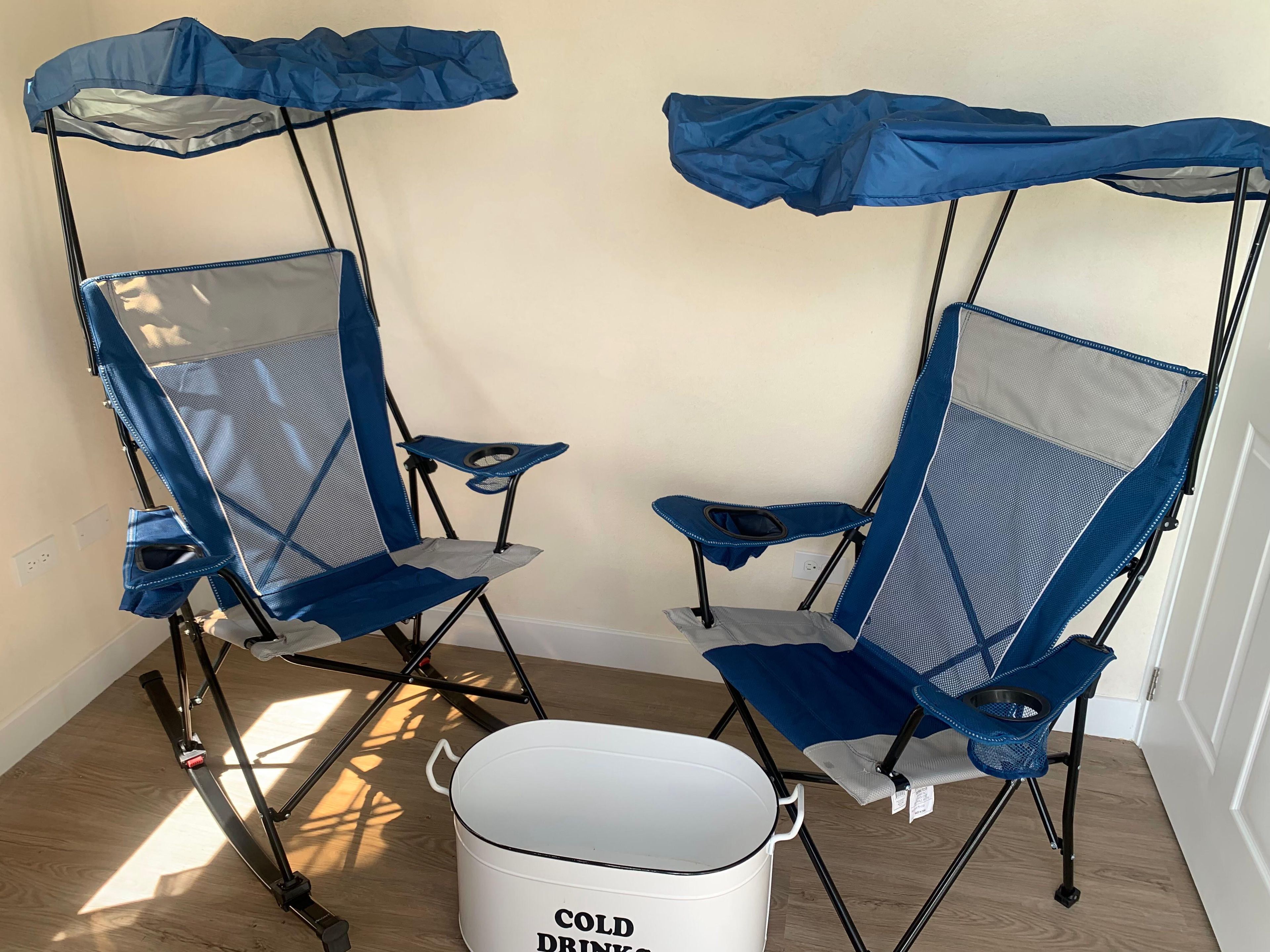 Two portable camping chairs with attached umbrellas, a white bucket labeled 'COLD DRINK,' and a wooden floor.