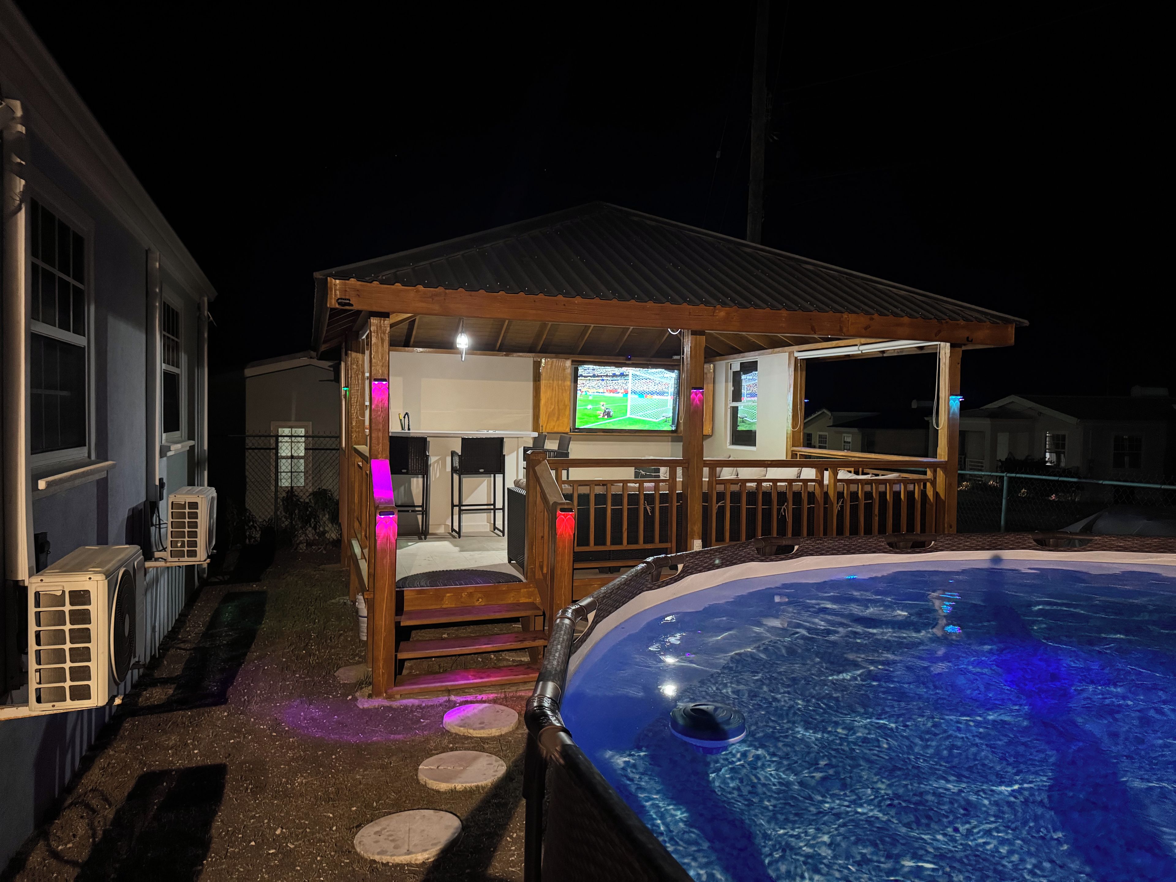 A pool area with a wooden deck, a bar area with a television, and a hot tub.