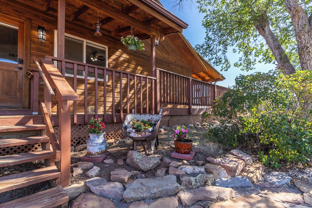 A wooden cabin with a porch, a wheelbarrow with flowers, and a potted plant.