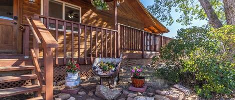 A wooden cabin with a porch, a wheelbarrow with flowers, and a potted plant.
