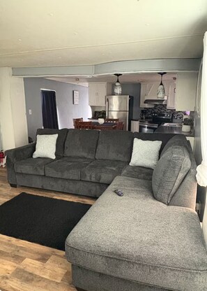 A living room with a grey sectional sofa, a black rug, and a kitchen area with a refrigerator and stove. - 3-bedroom mobile home in beautiful Eagle Pass with AC (Eagle Pass)