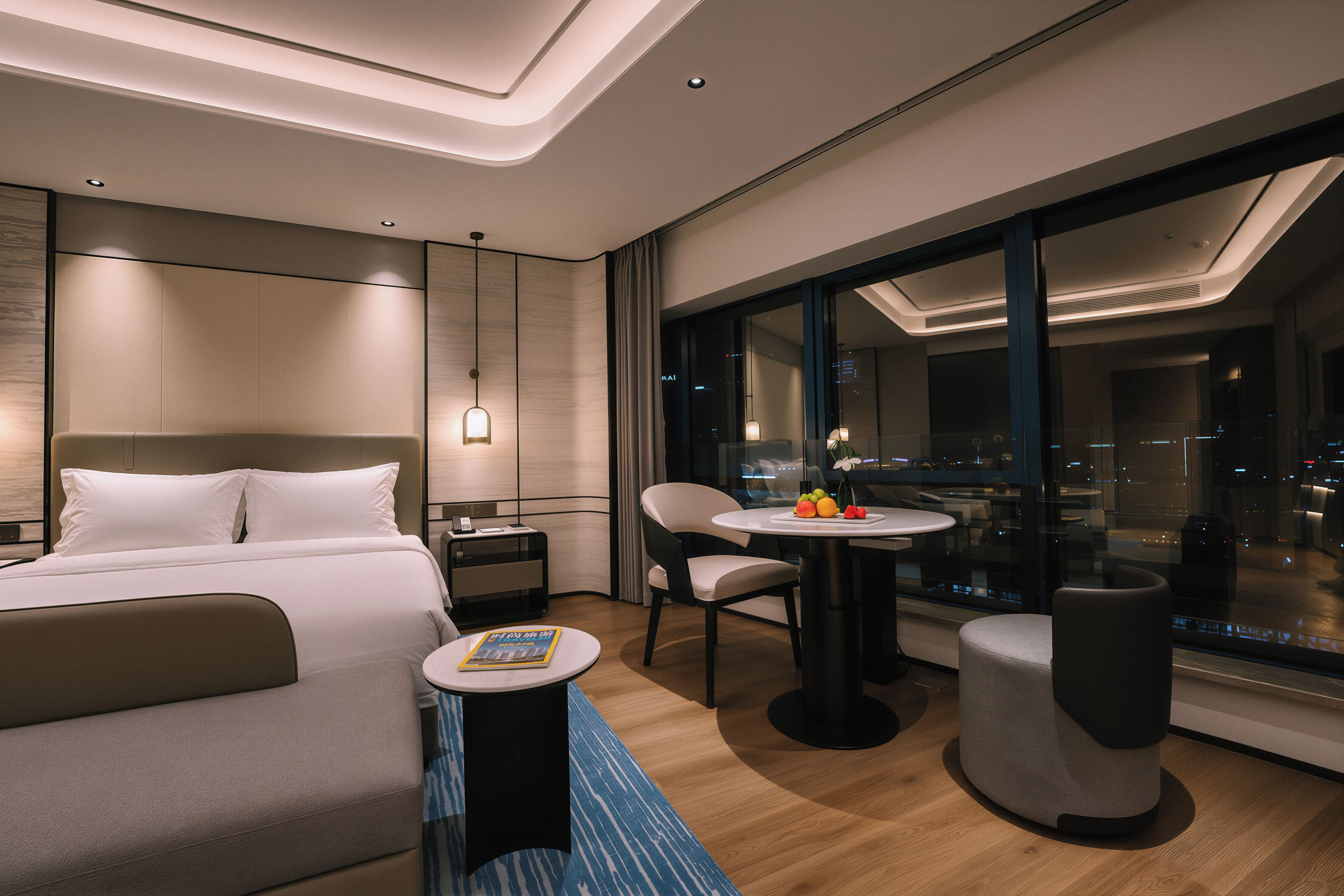 Une chambre d’hôtel moderne équipée d’un lit, d’un bureau, d’une chaise, d’une petite table avec des fruits, et offrant une vue sur la ville la nuit.