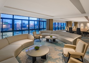 Una sala de conferencias moderna con vistas panorámicas de la ciudad, un sofá curvo, mesas redondas y una mesa de reuniones con sillas.