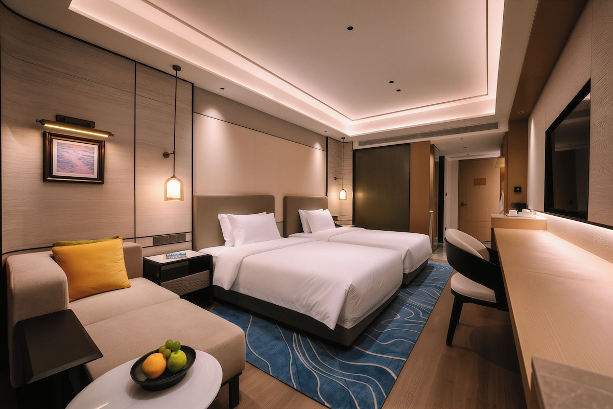 A modern hotel room with a large bed, a sofa, a desk, and a small table with fruit.