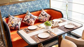 A dining area with a table set for two, a sofa with floral cushions, and a decorative vase with greenery.