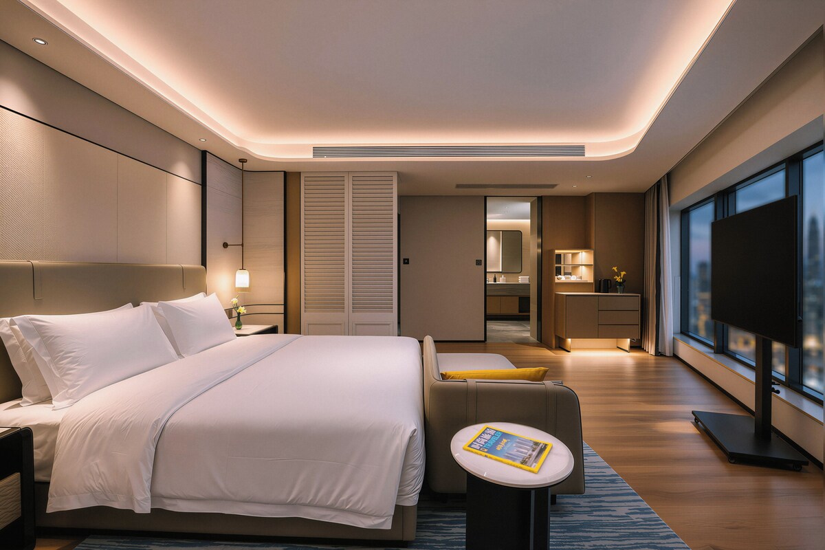 A modern hotel room with a large bed, a bedside table, a small sofa, a TV, and a view of the city through large windows.