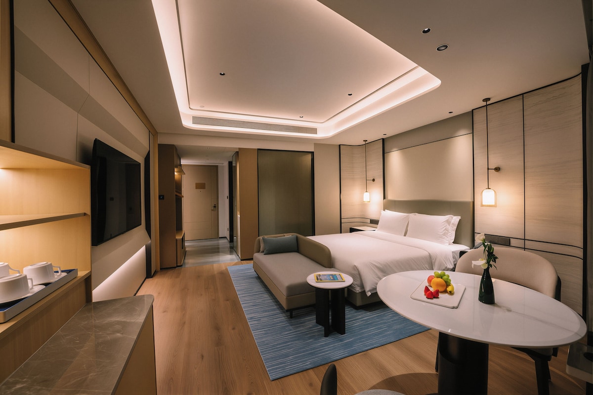 A modern hotel room with a bed, bedside table, and a small round table with fruit.