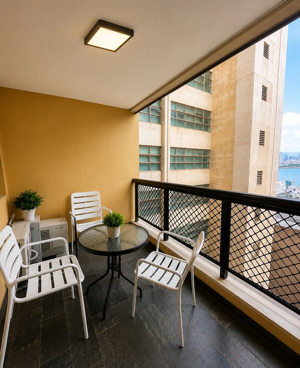Een balkon met een ronde tafel en vier stoelen, een bloempot en uitzicht op een stadslandschap achter een glazen balustrade.