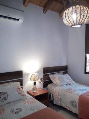 Un dormitorio con dos camas, una mesita de noche con lámpara, aire acondicionado e iluminación colgante.