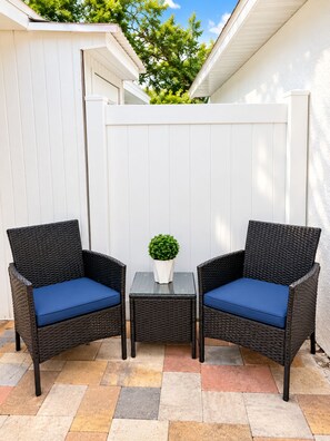 Zwei Korbstühle mit blauen Kissen und ein kleiner Tisch mit einem Blumentopf dazwischen, auf einer gefliesten Terrasse platziert.