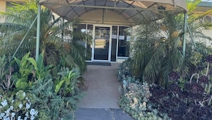 A covered entrance with a pathway leading to a building, surrounded by various plants and palm trees.