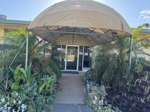 Un acceso cubierto con un sendero que lleva a un edificio, rodeado de plantas y palmeras.
