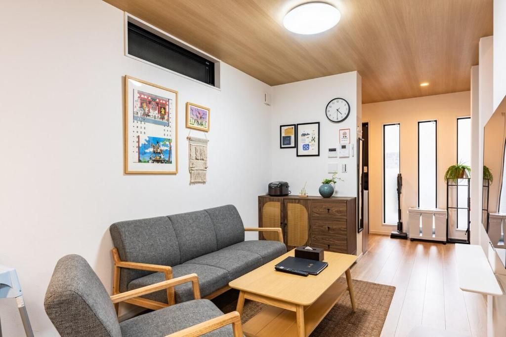 A modern living room with a grey sofa, wooden chairs, a coffee table, and a kitchen area with cabinets and a clock on the wall.