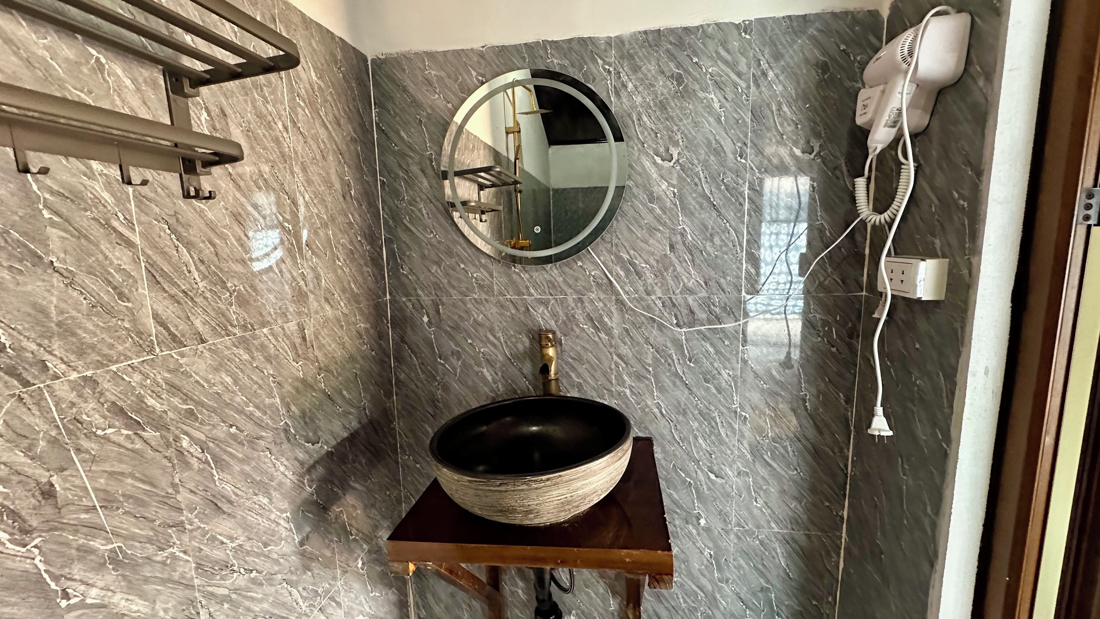Un baño con pared de mármol, un espejo redondo, un lavabo con plato, un perchero y un secador de pelo.