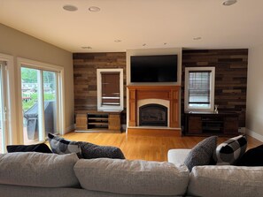 A living room with a fireplace, a television mounted above it, and a large window with blinds.