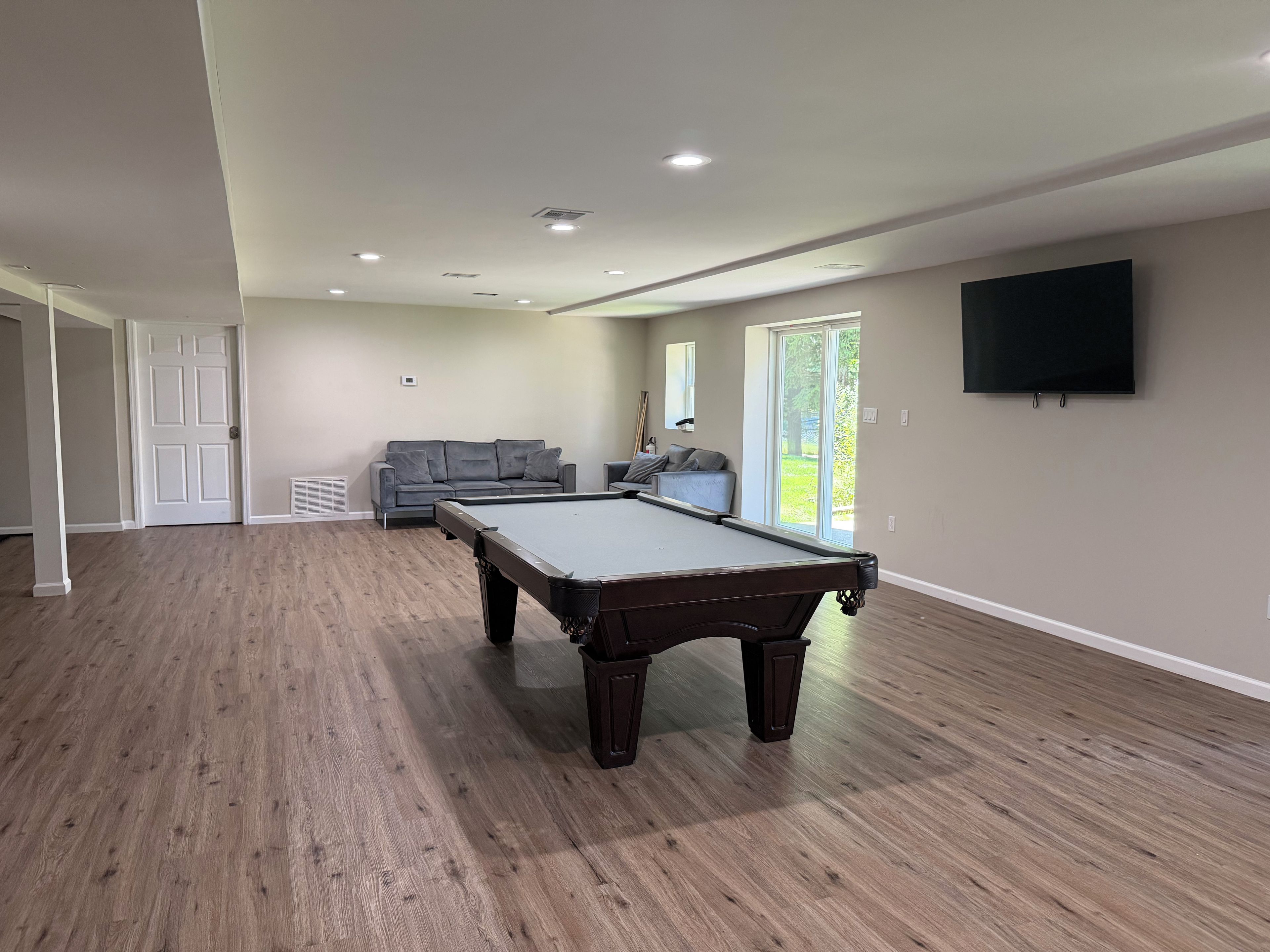 Una habitación amplia con mesa de billar, un sofá gris y un televisor montado en la pared.