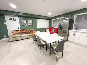Un comedor con una mesa y sillas blancas, un sofá con cojines de colores, un refrigerador y un ventanal con persianas.