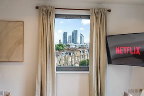Ein Zimmer mit Fensterblick auf eine Stadtlandschaft, einem Fernseher mit Netflix-Logo und einem gerahmten Bild an der Wand.