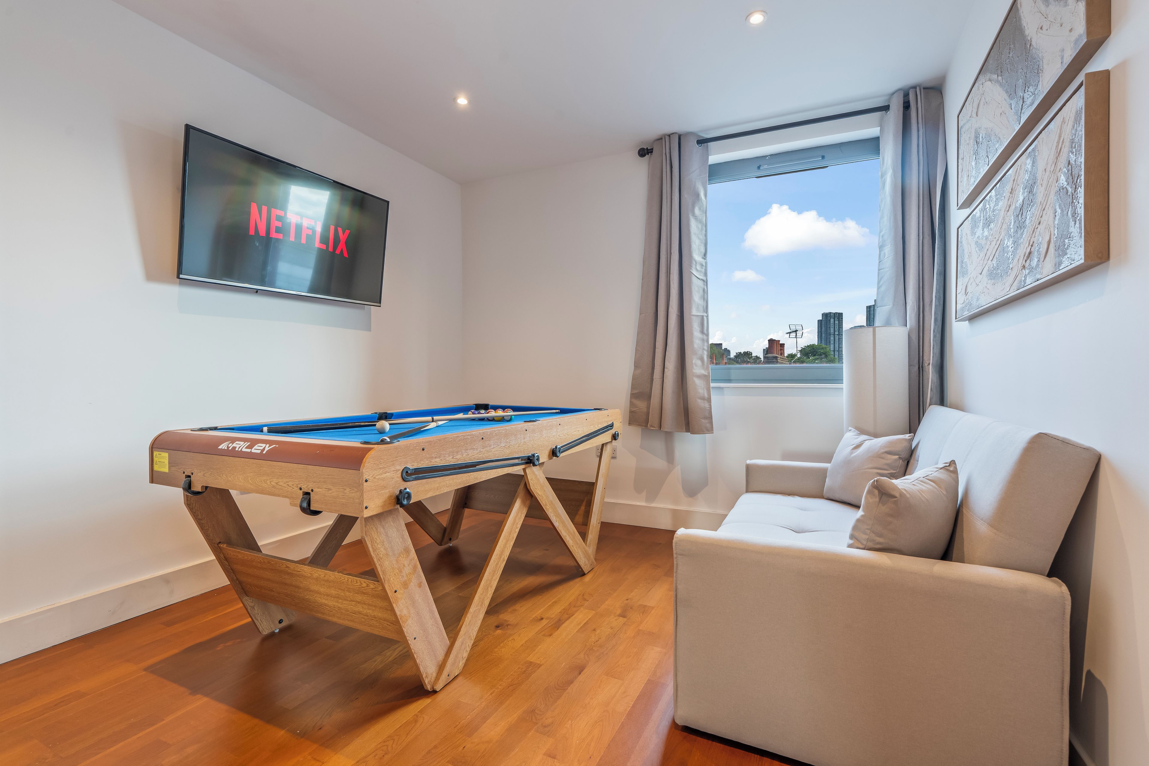 Una sala de estar moderna con mesa de billar, un sofá cómodo, un ventanal con cortinas y un televisor de pared mostrando 'NETFLIX'.