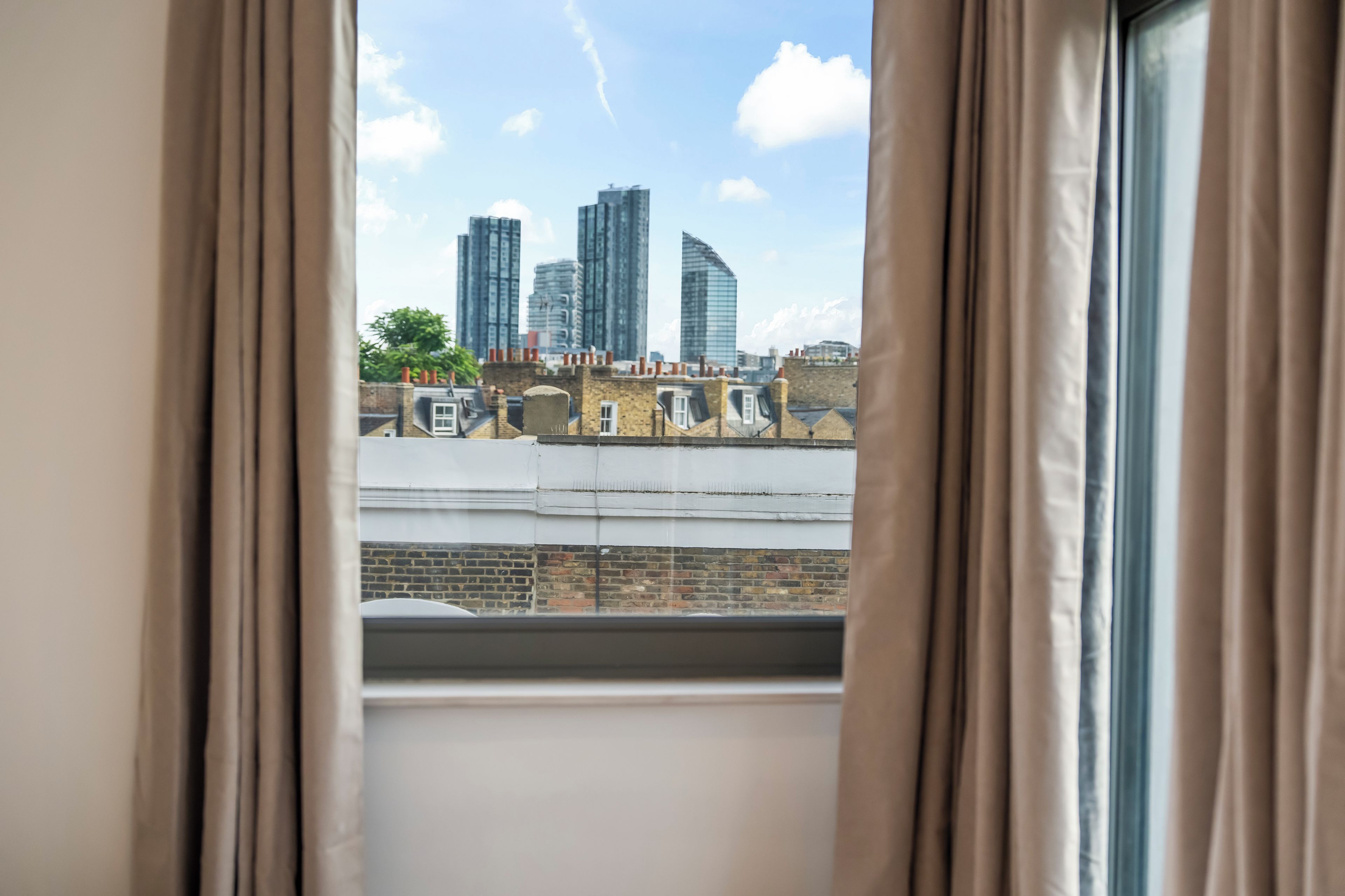 Une vue à travers une fenêtre avec des rideaux, donnant sur un paysage urbain avec des bâtiments modernes et des exemples d’architecture traditionnelle.
