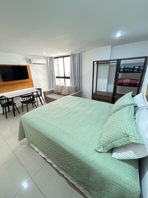A hotel room with a bed, a desk with chairs, a television, and a window with curtains. - Melhores Flats - Esplêndido, beira-mar (João Pessoa)