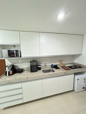 Eine Küche mit weißen Schränken, einer Arbeitsplatte, einer Kaffeemaschine, einem Wasserkocher, einem Toaster, einer Spüle und einem kleinen Kühlschrank.