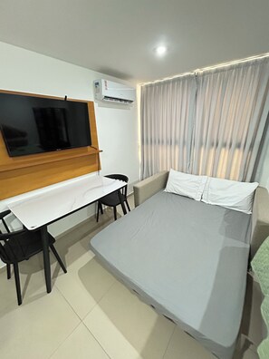 Ein Hotelzimmer mit einem Bett, einem an der Wand befestigten Fernseher, einem Schreibtisch mit Stuhl und einem Fenster mit Vorhängen.