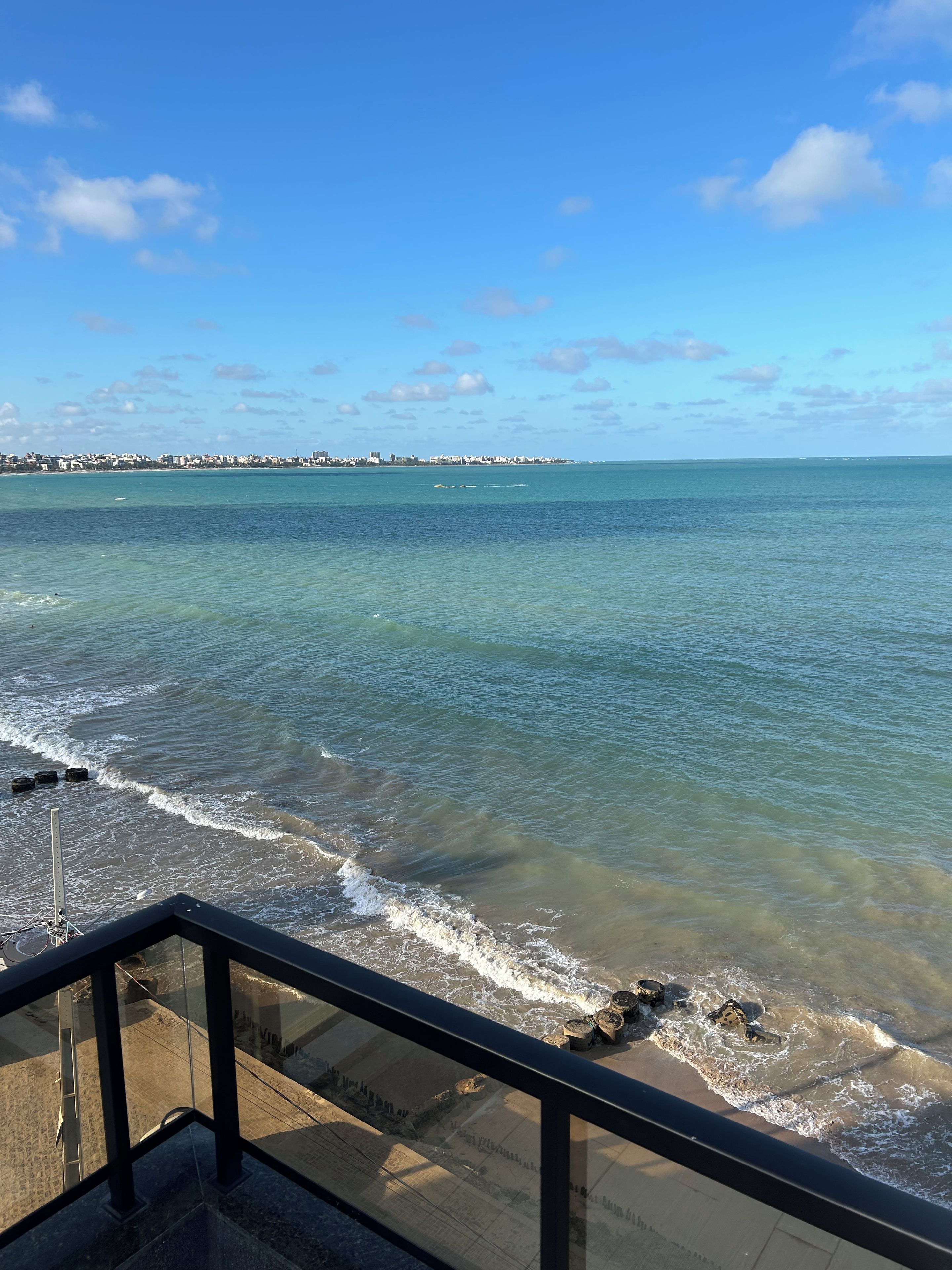 A coastal view from a balcony with a clear sky, ocean waves, and distant buildings.