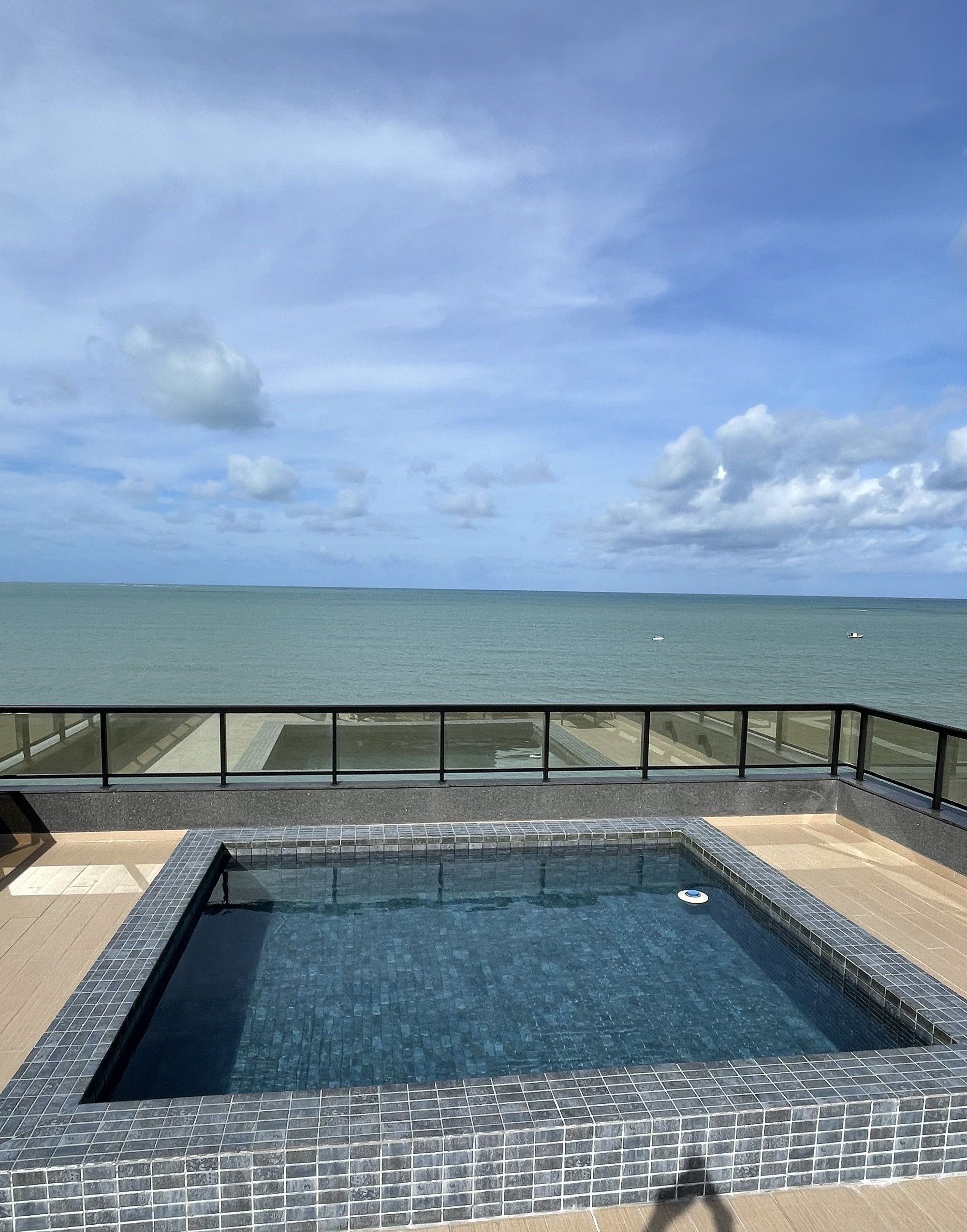 A rectangular pool with tiled edges, surrounded by a glass railing, overlooking a calm sea under a partly cloudy sky.