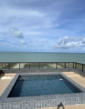 Ein rechteckiger Pool mit Fliesenrand, umgeben von einem Glasgeländer, mit Blick auf ein ruhiges Meer unter einem teils bewölkten Himmel.