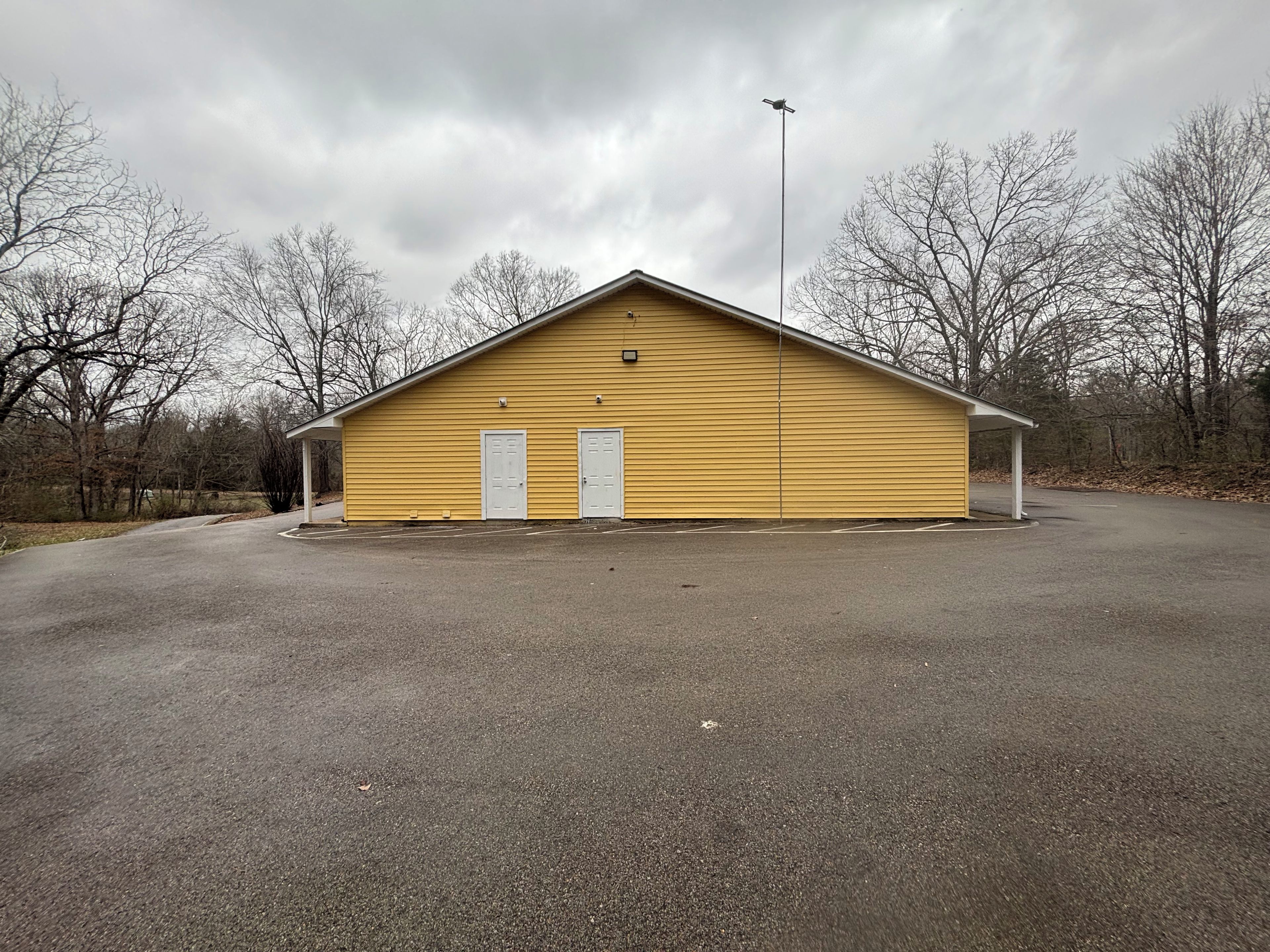 A yellow building with two doors and a covered area, surrounded by a paved area and leafless trees.