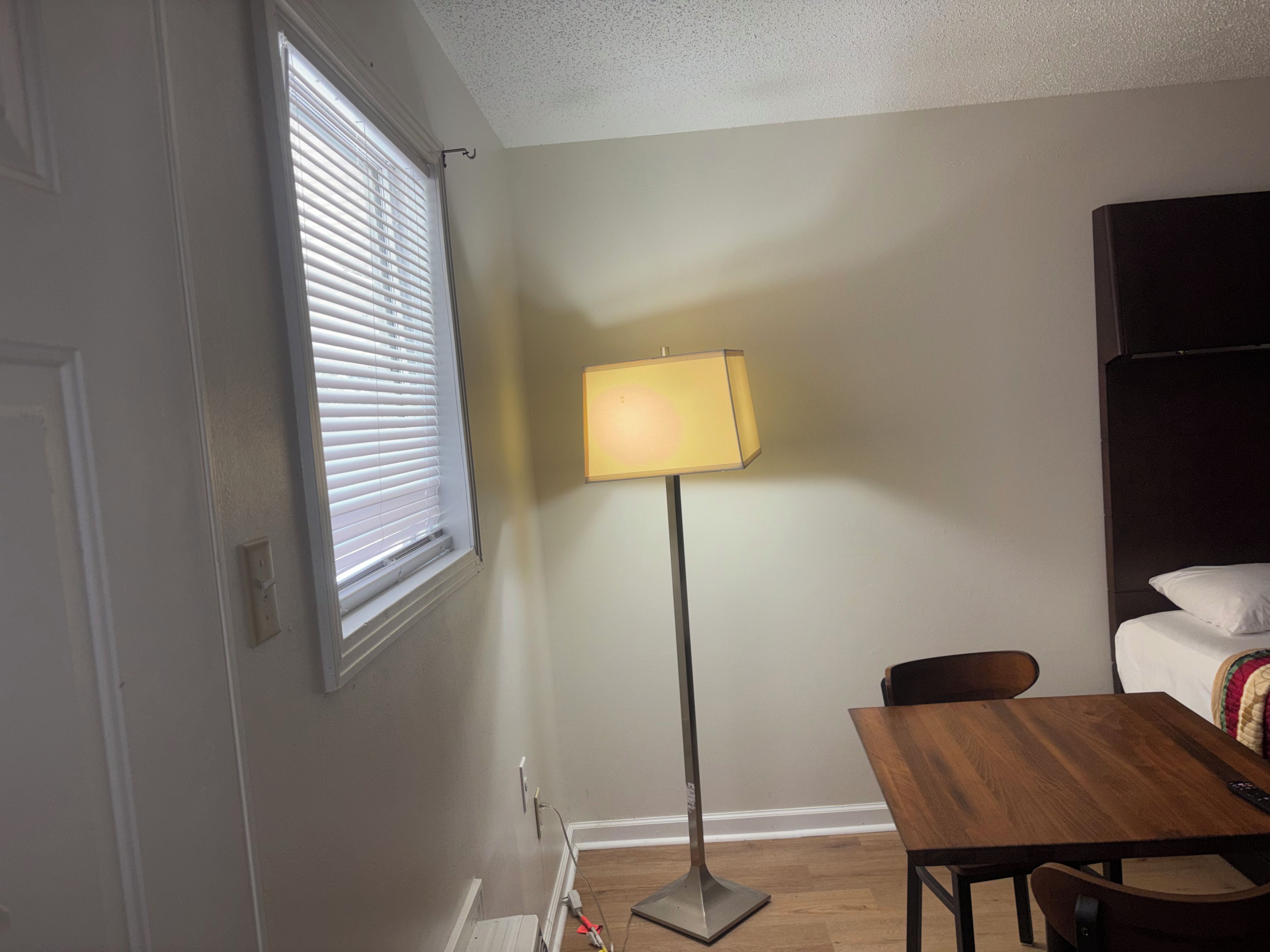 A room with a window, a standing floor lamp, a wooden table, and a bed with a white pillow.