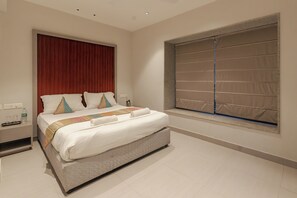 A hotel room with a bed, a nightstand, a window with blinds, and a wall-mounted outlet.
