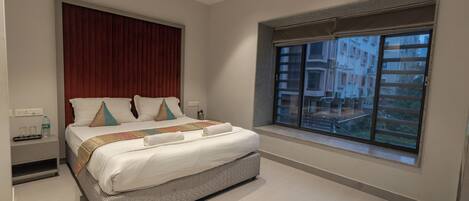 A modern hotel room with a large bed, a window with a view of buildings, and a small bedside table with a bottle and glasses.