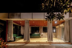 An indoor-outdoor living space with a wooden ceiling, green seating, and curtains.