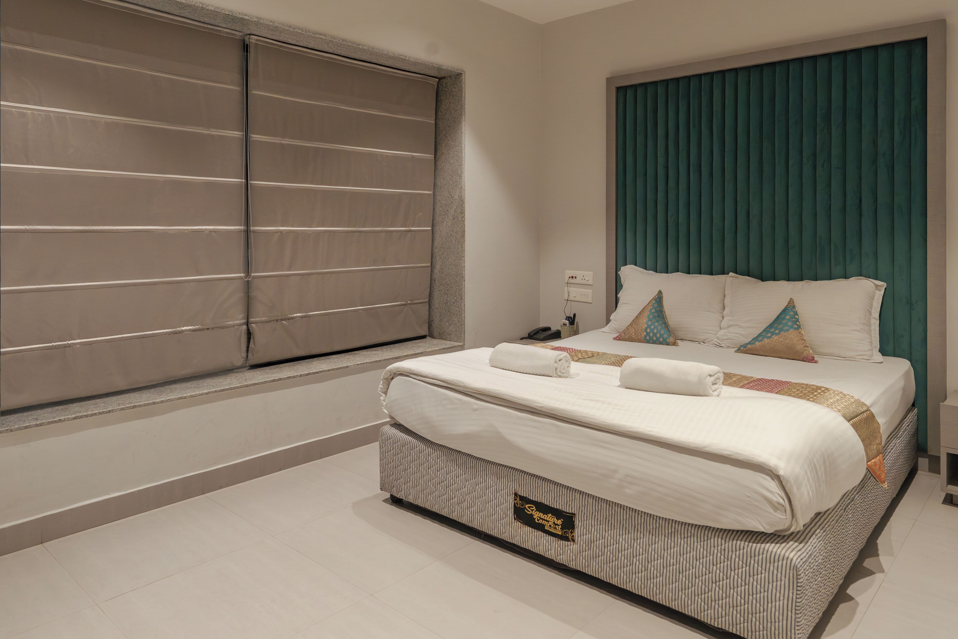 Habitación de hotel con una cama grande, una ventana con persianas, una mesita de noche con lámpara y un cabecero con cortinas.