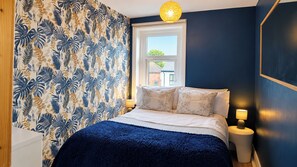 Une chambre à coucher avec un lit, deux lampes de chevet, une fenêtre et un mur orné d’un motif de feuilles tropicales.