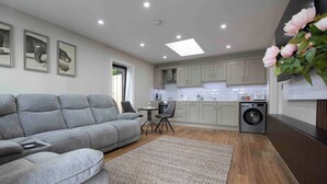 A modern kitchen with grey cabinets, a built-in washing machine, and a grey sofa in the living area. - Meigh Village Apartment (Newry)
