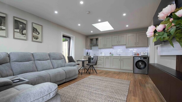 A modern kitchen with grey cabinets, a built-in washing machine, and a grey sofa in the living area. - Meigh Village Apartment (Newry)