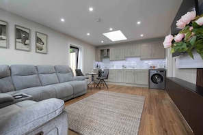 A modern kitchen with grey cabinets, a built-in washing machine, and a grey sofa in the living area. - Meigh Village Apartment (Newry)